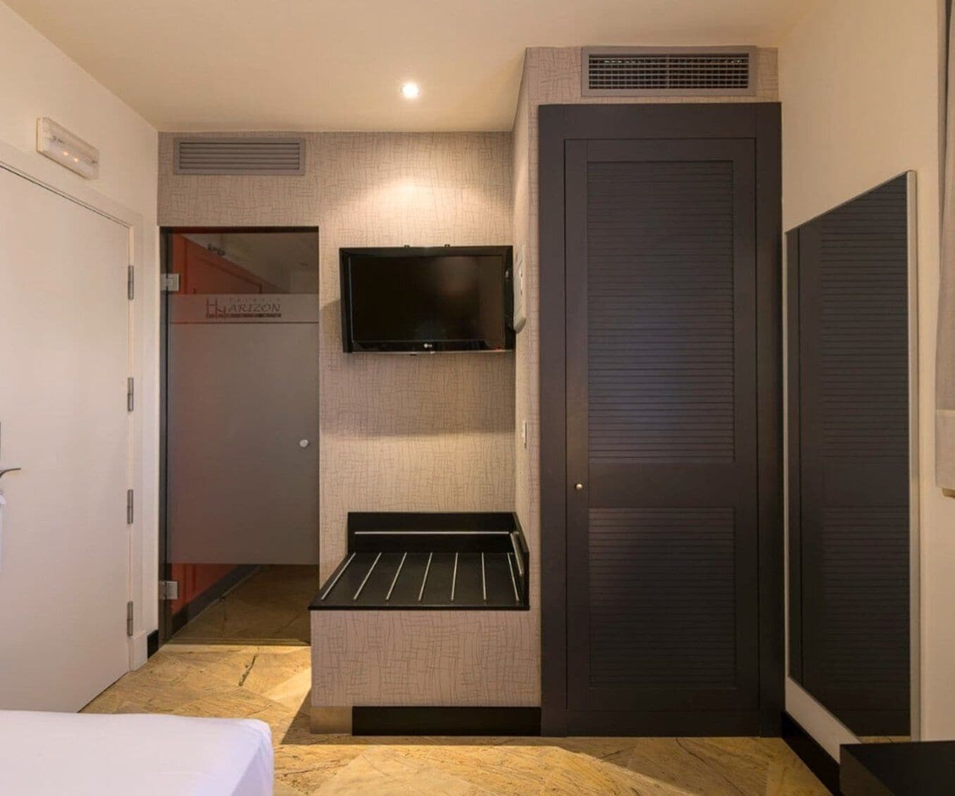 Habitación de hotel compacta con cama, televisión de pantalla plana en la pared, un pequeño espacio de descanso y un armario.