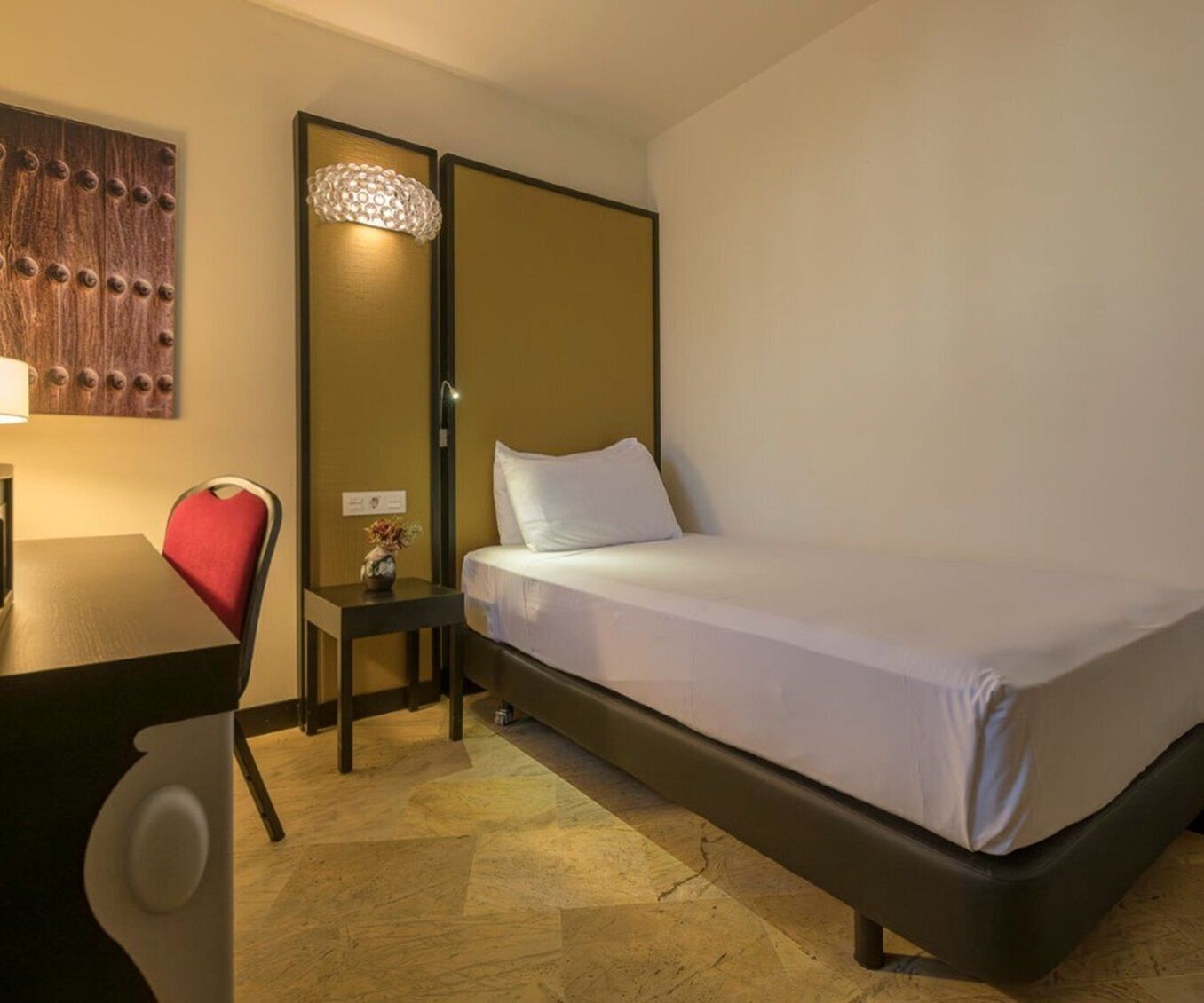 Habitación de hotel con una cama individual, un escritorio con silla, una mesita con un jarrón y una lámpara fijada a la pared.
