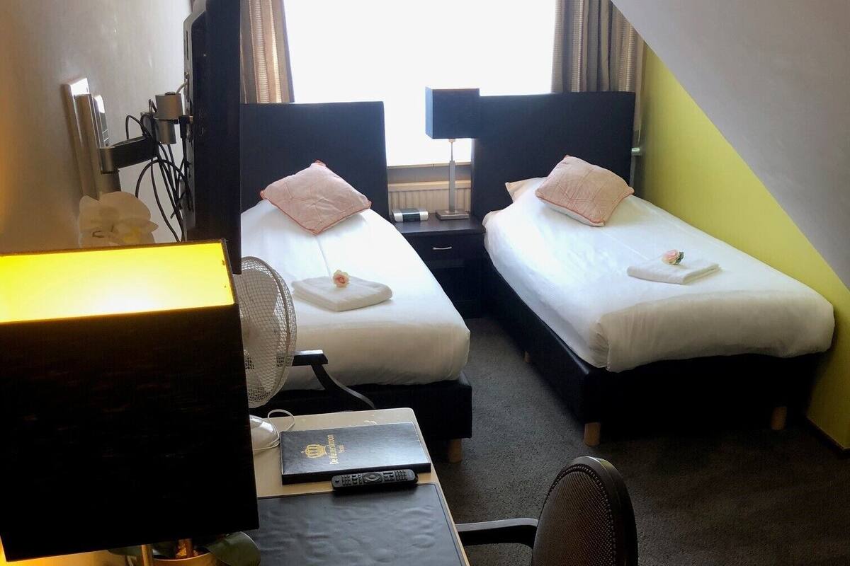 Een hotelkamer met twee éénpersoonsbedden, een bureau met een laptop, een stoel, een bureaulamp, een ventilator en een raam met gordijnen.