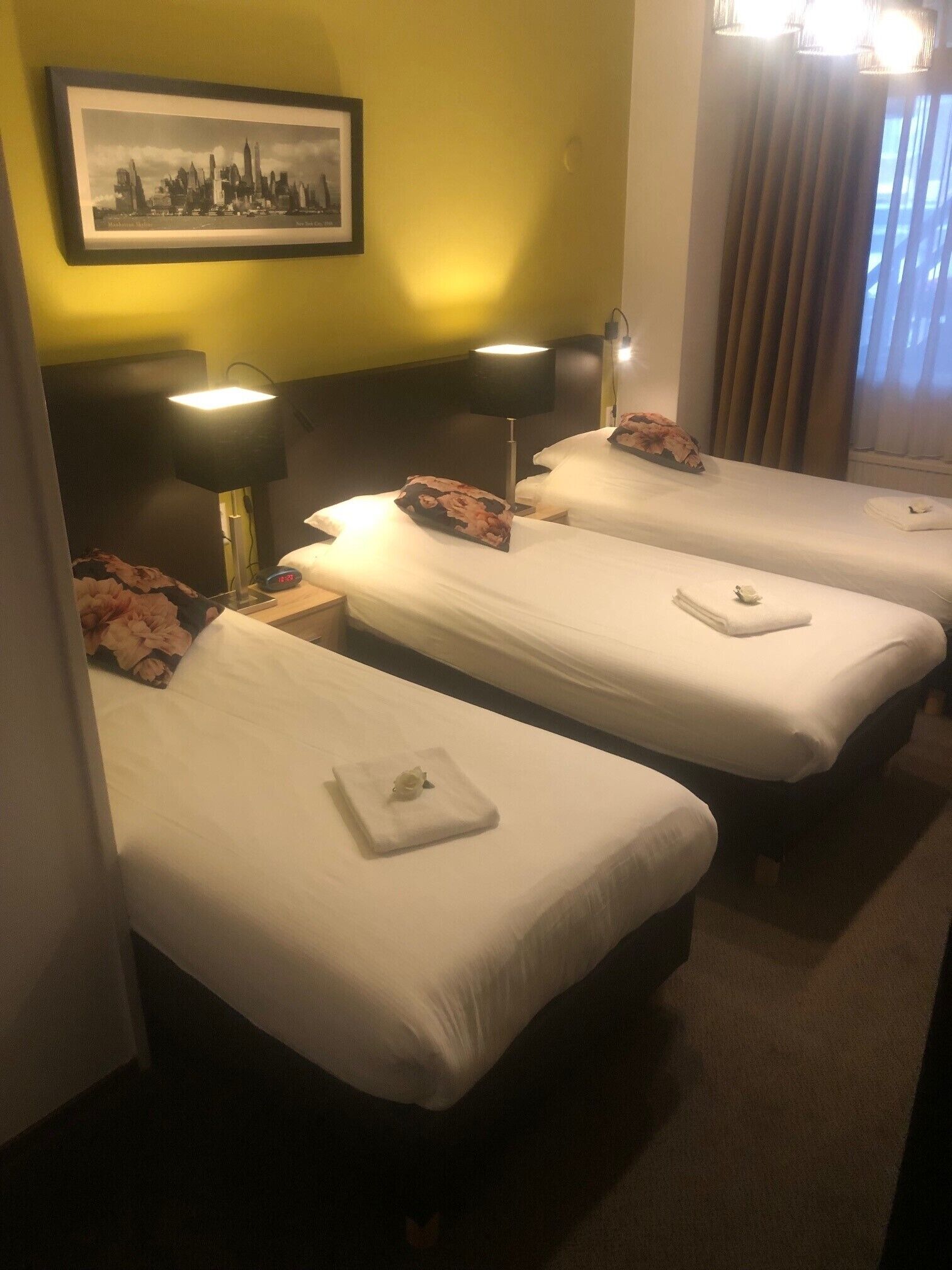 A hotel room with three single beds, each with a pillow and a folded towel. There is a framed picture on the wall above the beds, and a lamp on each bedside table.