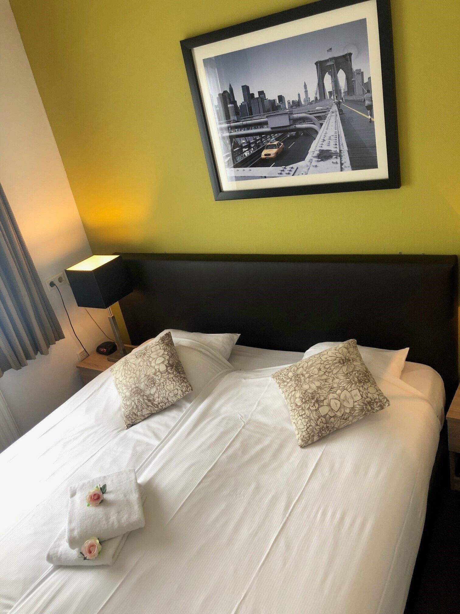 Un lit bien fait, agrémenté de coussins à motifs floraux, une photographie encadrée représentant un paysage urbain accrochée au mur et une lampe de chevet.