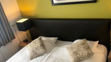 Un lit bien fait, agrémenté de coussins à motifs floraux, une photographie encadrée représentant un paysage urbain accrochée au mur et une lampe de chevet.