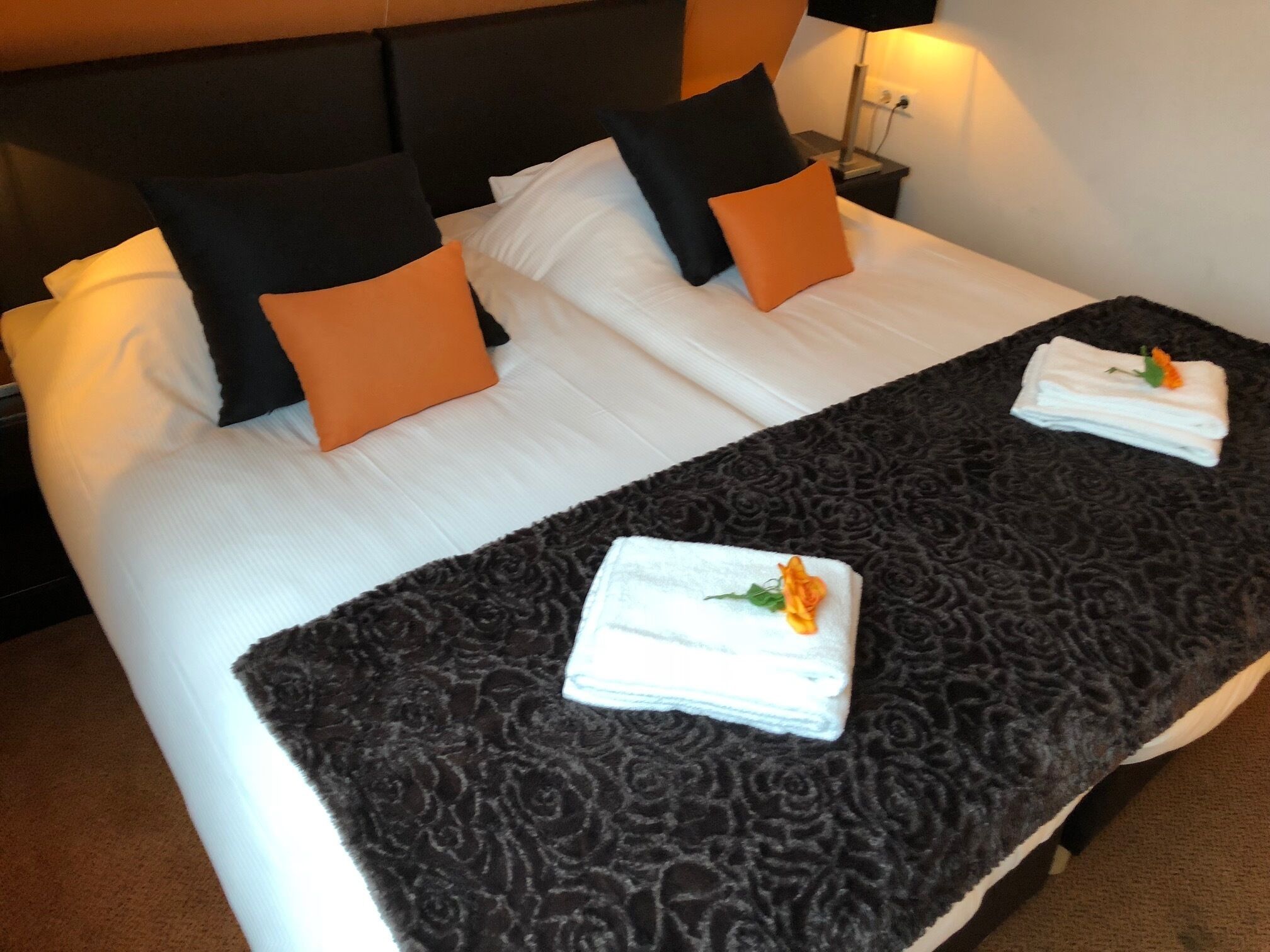Una cama bien hecha con un edredón oscuro estampado, sábanas blancas y almohadas naranjas.