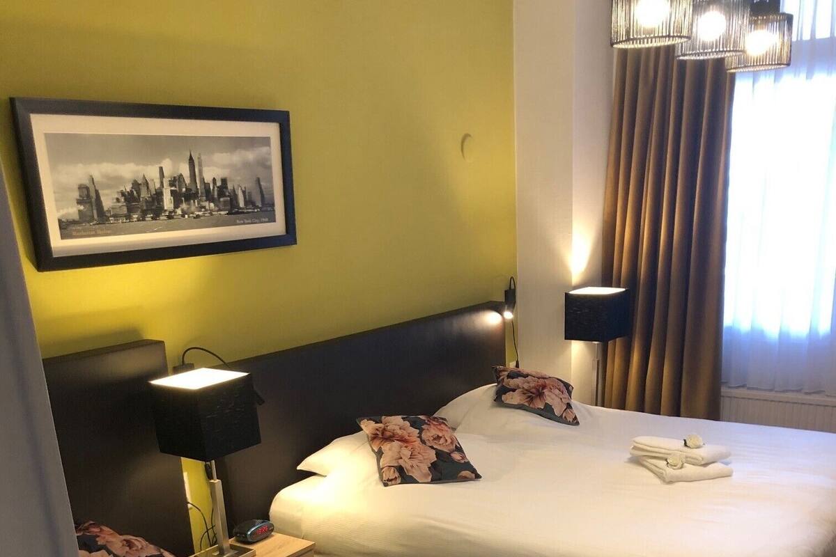 Hotelkamer met twee bedden, een nachtkastje met een lamp, een stadsgezicht in een lijst aan de muur, en een raam met gordijnen.