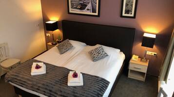 A hotel room with a large bed, two pillows, a bedside table with a lamp, a chair, and two framed pictures on the wall.