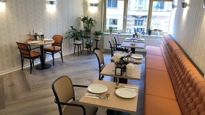 A modern dining area with wooden tables and chairs, a long orange cushioned bench, and potted plants. - Hotel De Keizerskroon (Hoorn)