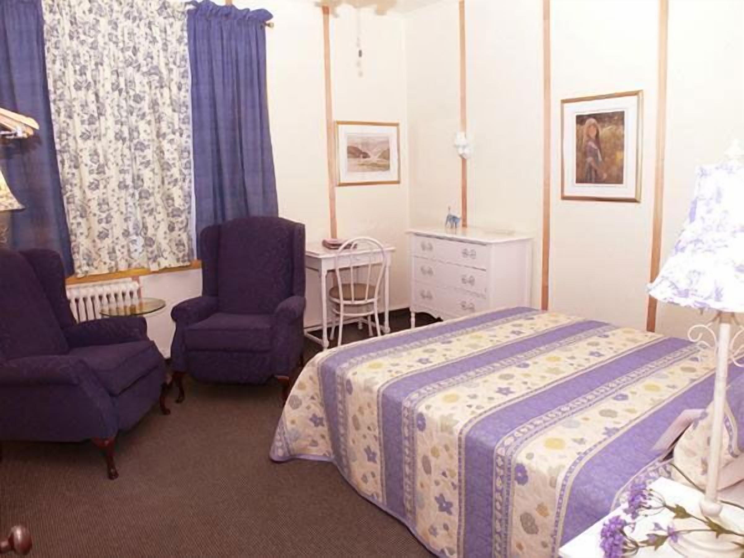 A bedroom with a bed, two chairs, a desk, a dresser, and a lamp.
