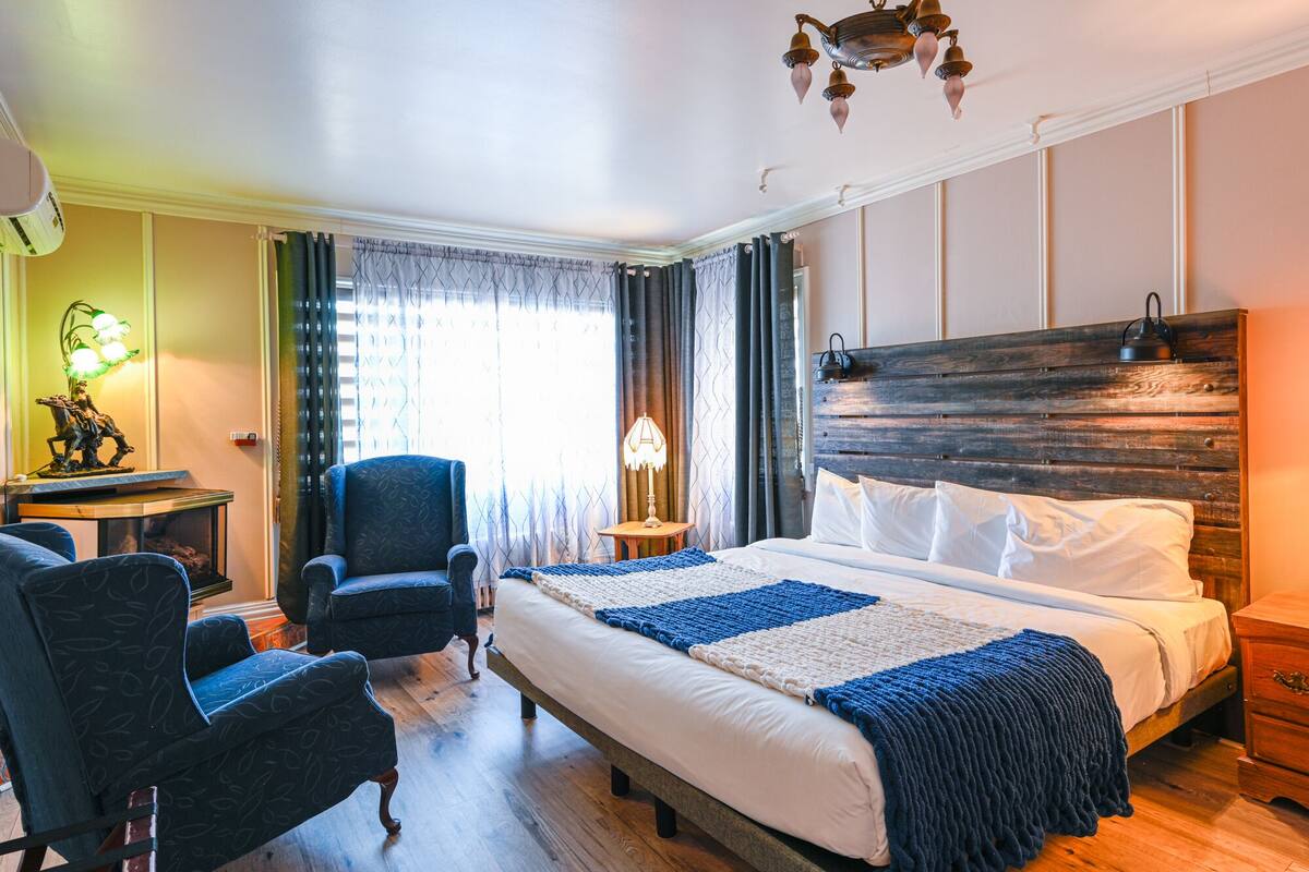 A hotel room with a large bed, two armchairs, a wooden headboard, a nightstand, and a lamp.