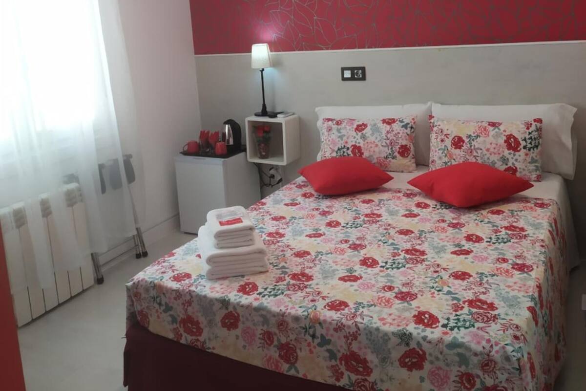 A bedroom with a floral bedspread, red pillows, a bedside table with a lamp, and a radiator.