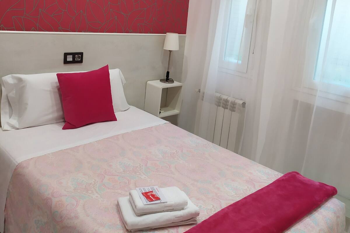 A hotel room with a bed, a nightstand, an air conditioning unit, a window with curtains, and a radiator.