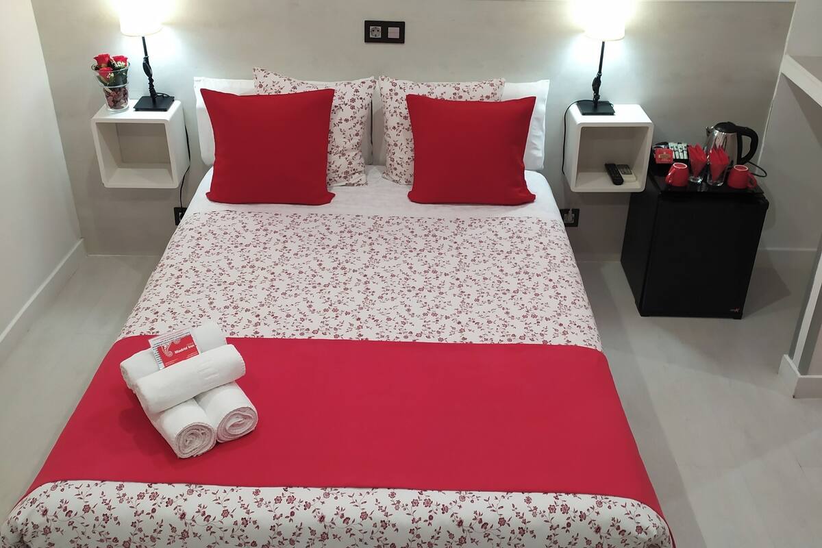 A neatly made bed with red and white floral bedding, two red pillows, and a set of white towels on the bed.