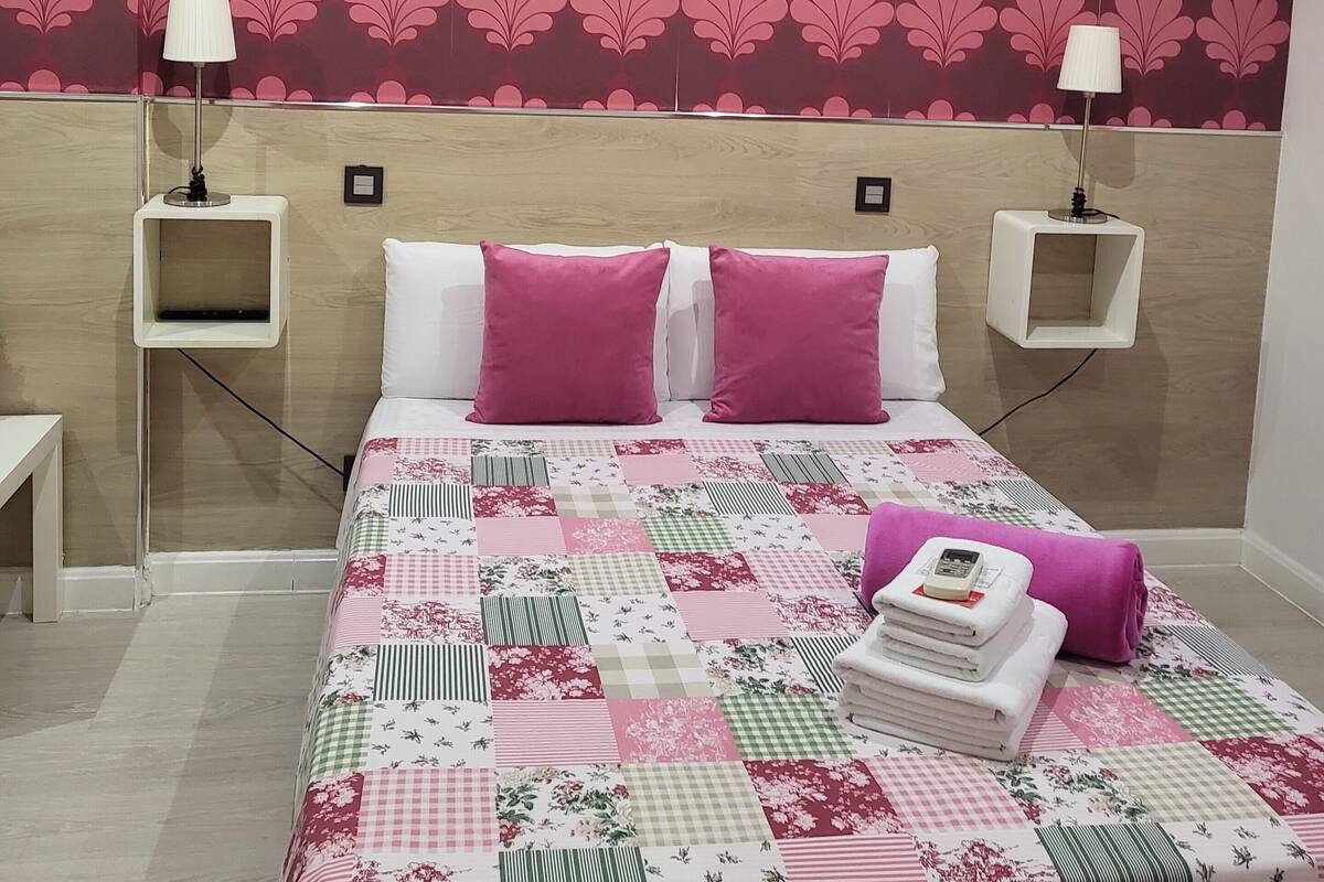 A neatly arranged bedroom with a floral-patterned headboard, two bedside lamps, a quilted bedspread, and a neatly folded towel on the bed.