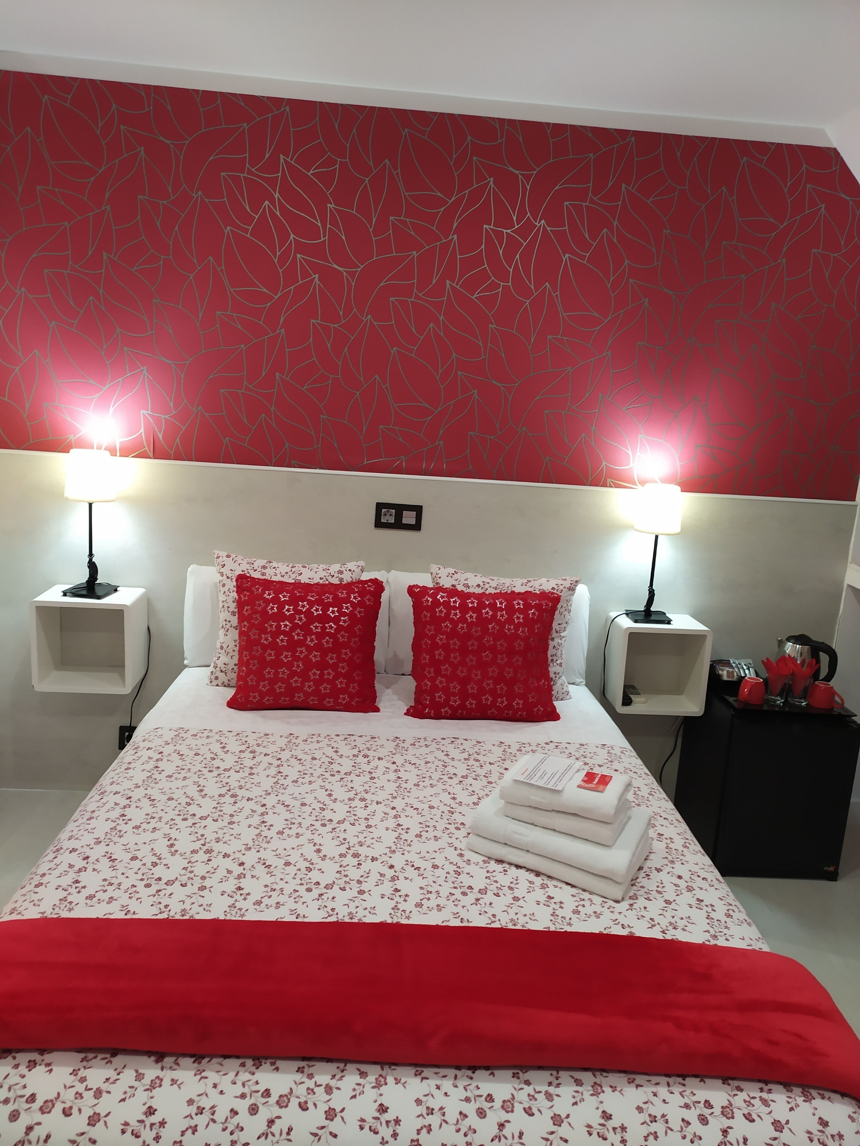 A hotel room with a red leaf-patterned wall, two bedside lamps, a bed with floral bedding, two red pillows, a red throw blanket, and a small black nightstand with red cups and a kettle.