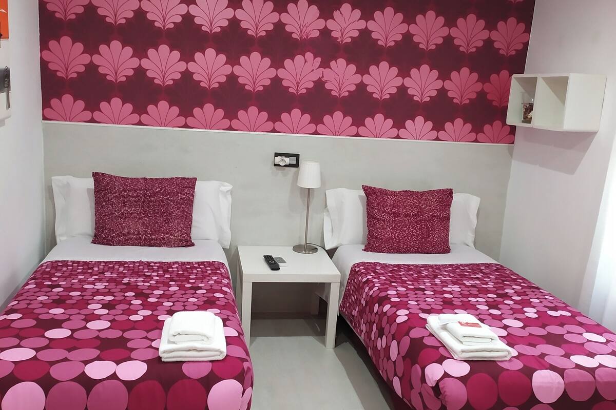 A hotel room with two beds, each with a red and pink polka dot bedspread, white pillows, and a red throw pillow. There is a nightstand with a lamp, a remote control, and a small shelf with a decorative item. The wall behind the beds has a large floral wallpaper.