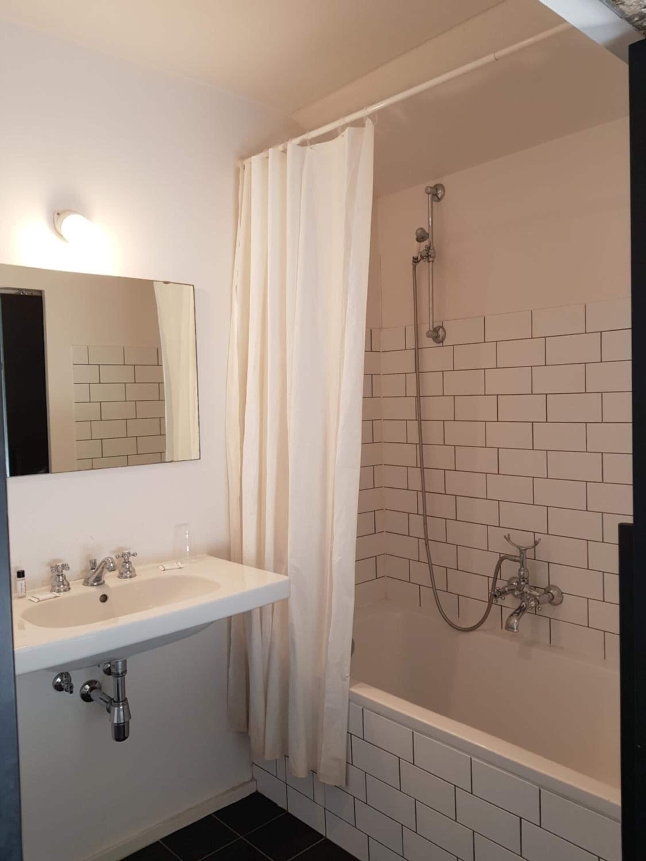 Un baño con un lavamanos blanco, un espejo, una ducha con cortina y paredes de azulejos.