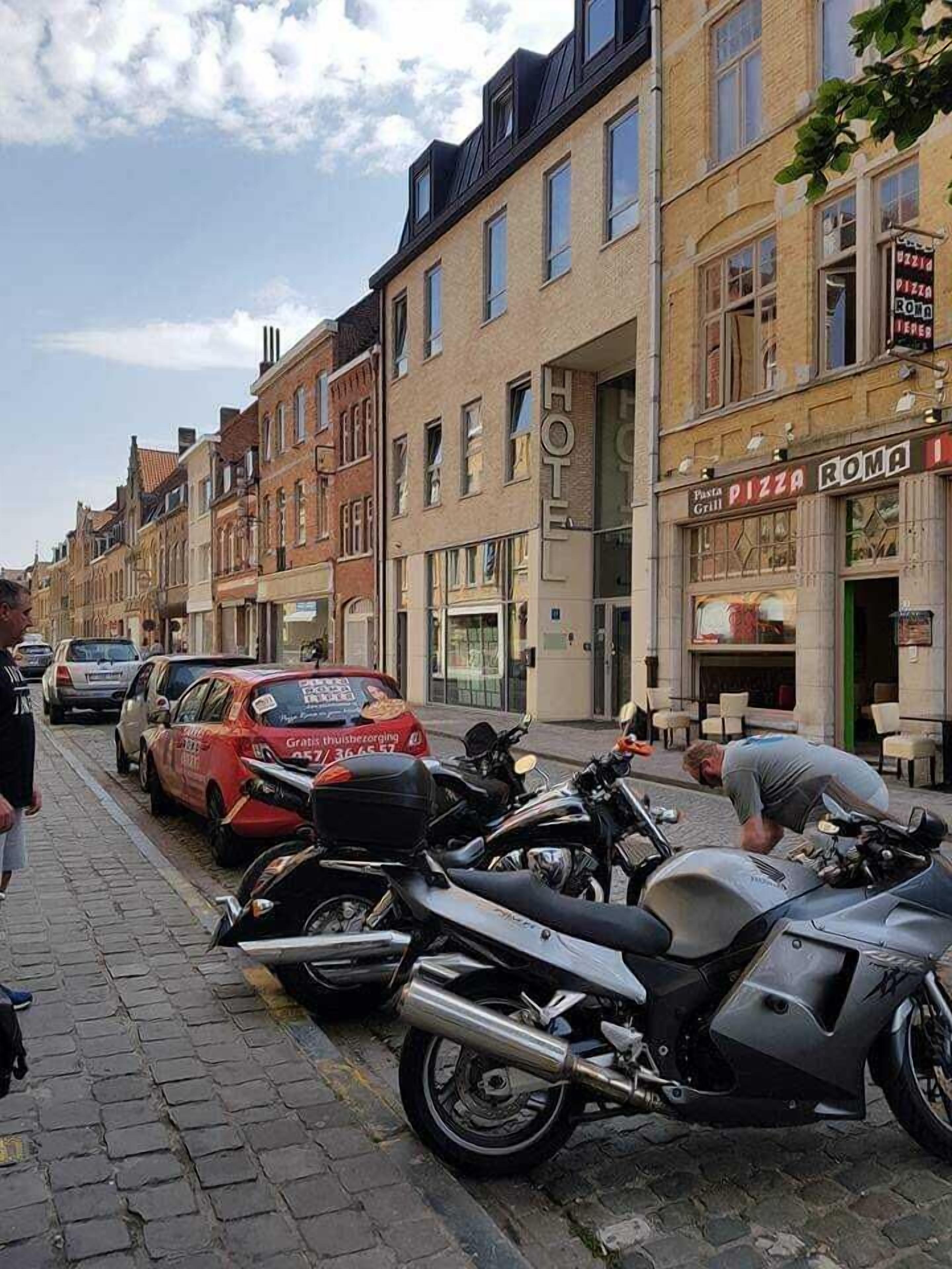 Une rangée de motos garées dans une rue pavée, un taxi rouge et un bâtiment sur lequel on peut lire l’inscription « PIZZA ROMA ».