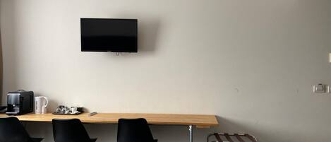 Una habitación de hotel moderna con un escritorio de madera, tres sillas negras, un televisor montado en la pared y un pequeño cubo de basura.