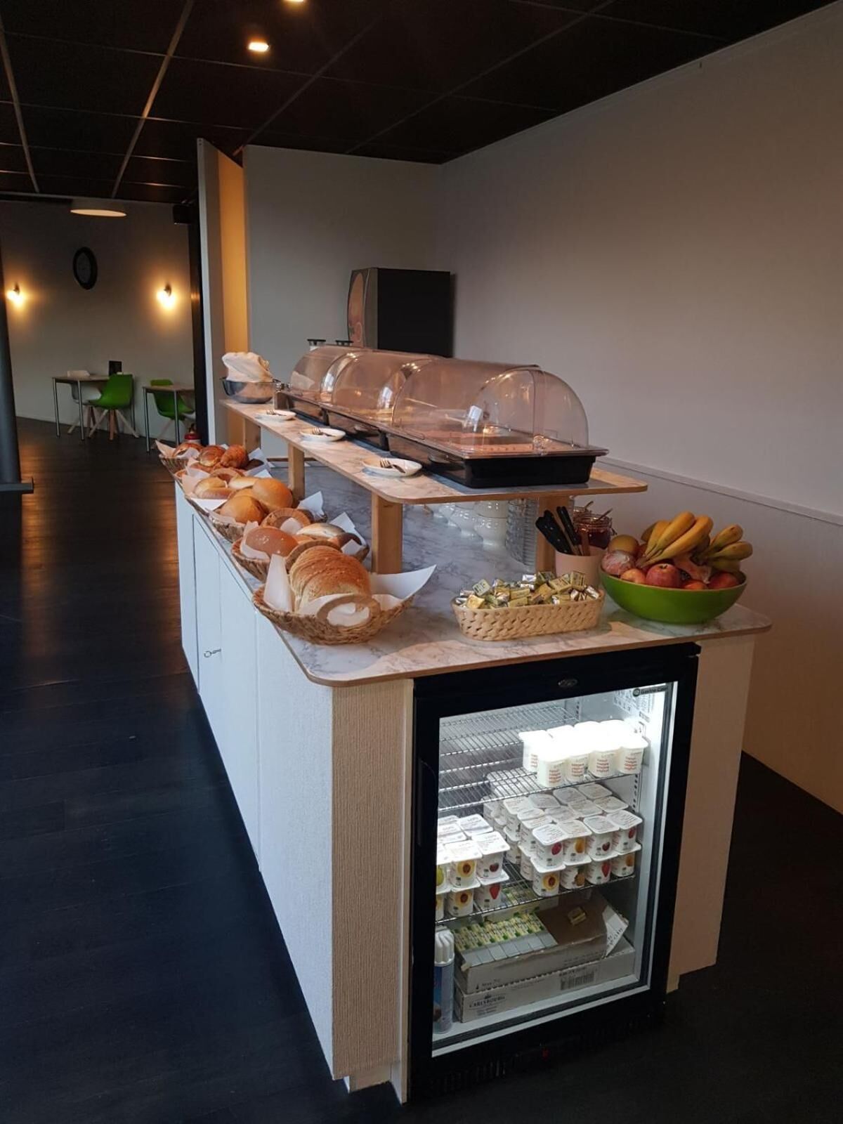Una barra de desayuno con pan, bollería y un bol de fruta, además de un refrigerador lleno de bebidas.
