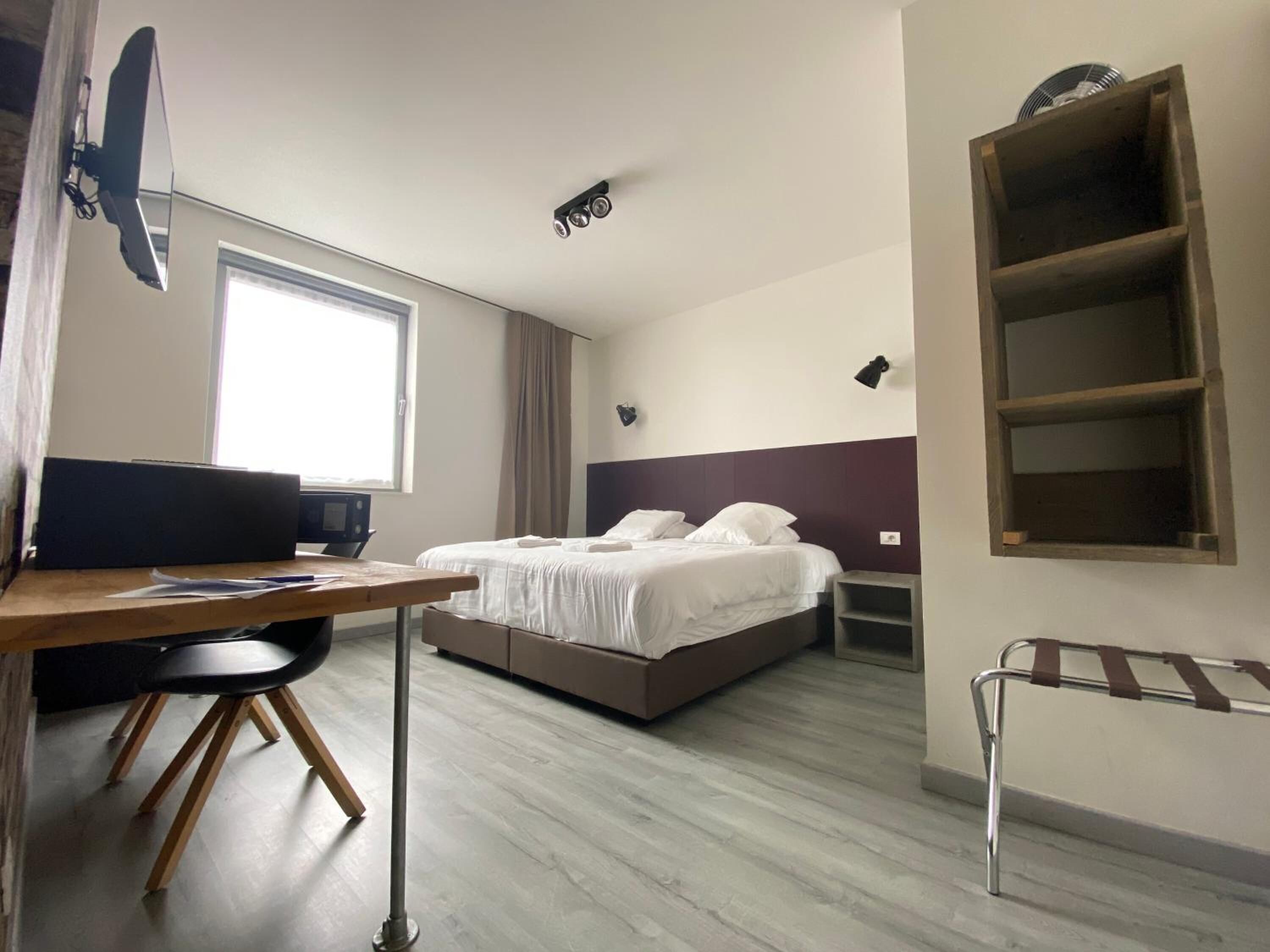 A hotel room with a bed, a desk with a computer, a chair, a window, and a shelf unit.