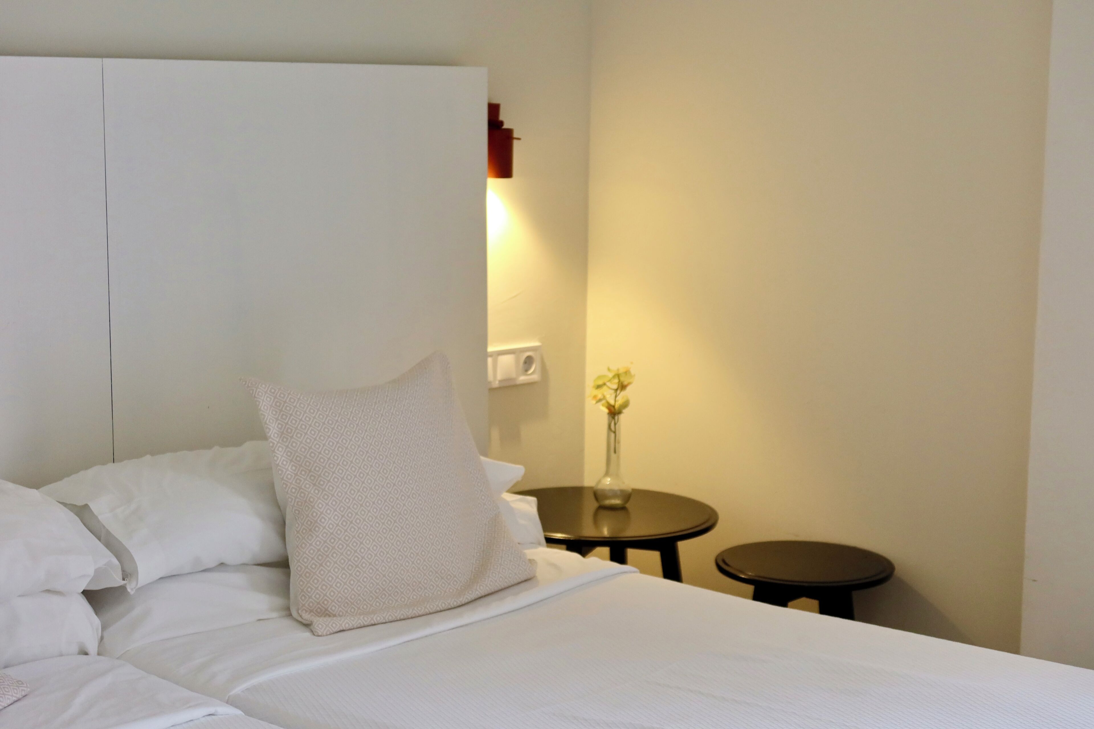 Een net opgemaakt bed met witte lakens en een gekleurd kussen, een nachtkastje met een bloevas, en een wandlamp.