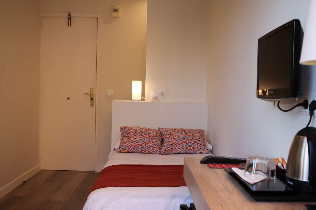 A hotel room with a bed, a TV mounted on the wall, a bedside table with a glass and a kettle, and a door.