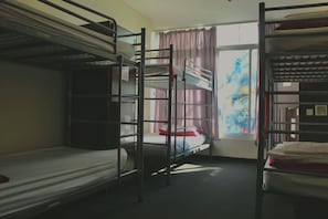 A dormitory room with bunk beds, a window with curtains, and a view of a building outside.