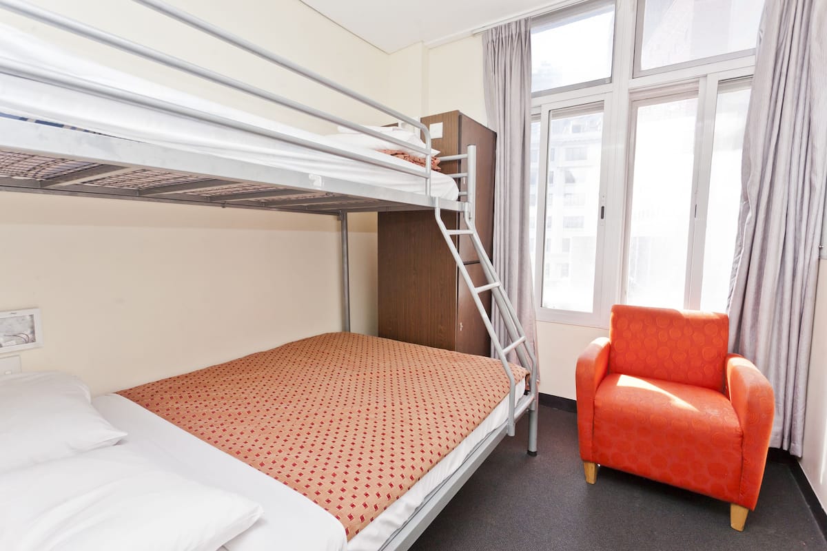 Un lit superposé avec une échelle, un lit simple et un fauteuil rouge dans une pièce dotée d’une fenêtre et de rideaux.