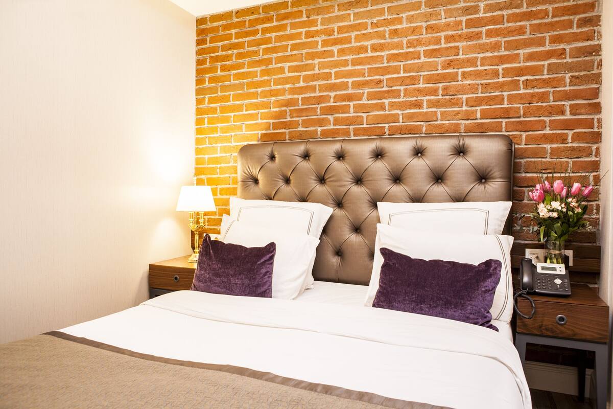 A hotel room with a brick wall, a tufted headboard, a bed with white linens and purple pillows, a nightstand with a lamp, and a vase with flowers.