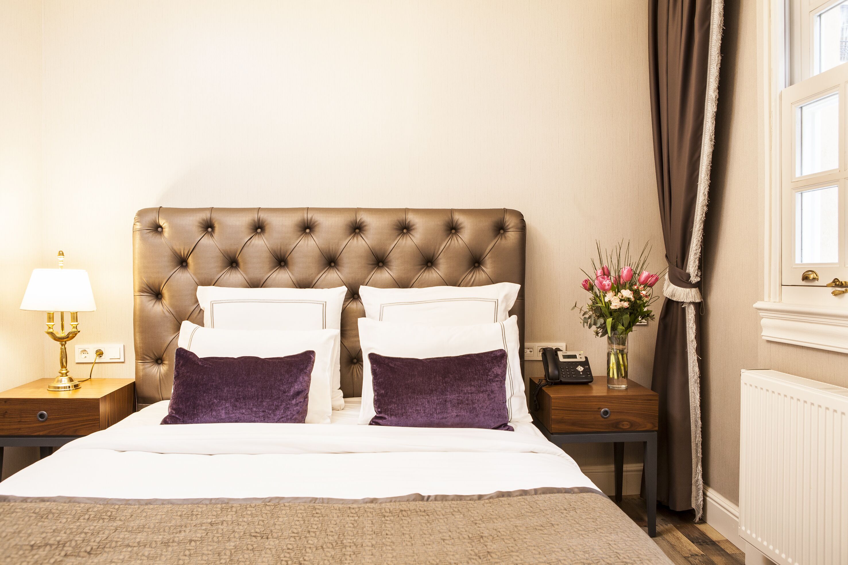 A hotel room with a tufted headboard, white bedding, purple pillows, a bedside table with a lamp, a vase with flowers, and a telephone.