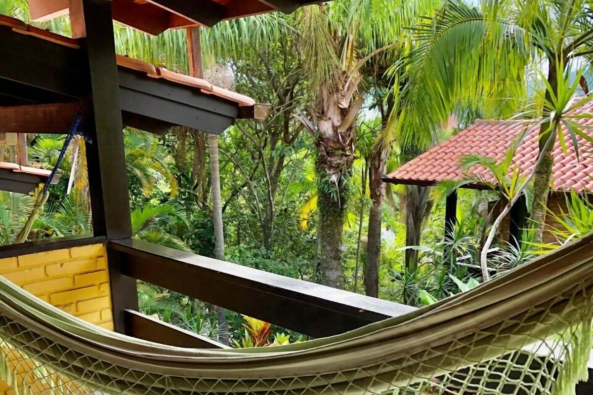 A hammock under a wooden pergola with a tiled floor, surrounded by lush greenery and a red-roofed structure.