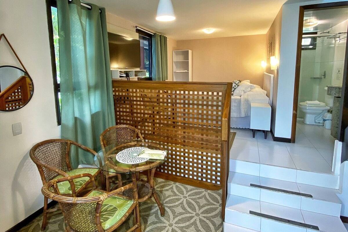 A hotel room with a dining area featuring wicker chairs and a glass table, a wooden partition, and a view of the bathroom.