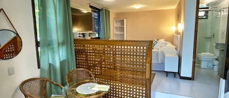 Habitación de hotel con comedor que incluye sillas de mimbre, una mesa de vidrio, un separador de madera y vistas al baño.