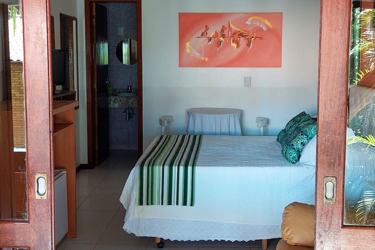 A bedroom with a bed, a bedside table, a chair, a hanging light fixture, and a bathroom visible through an open door.