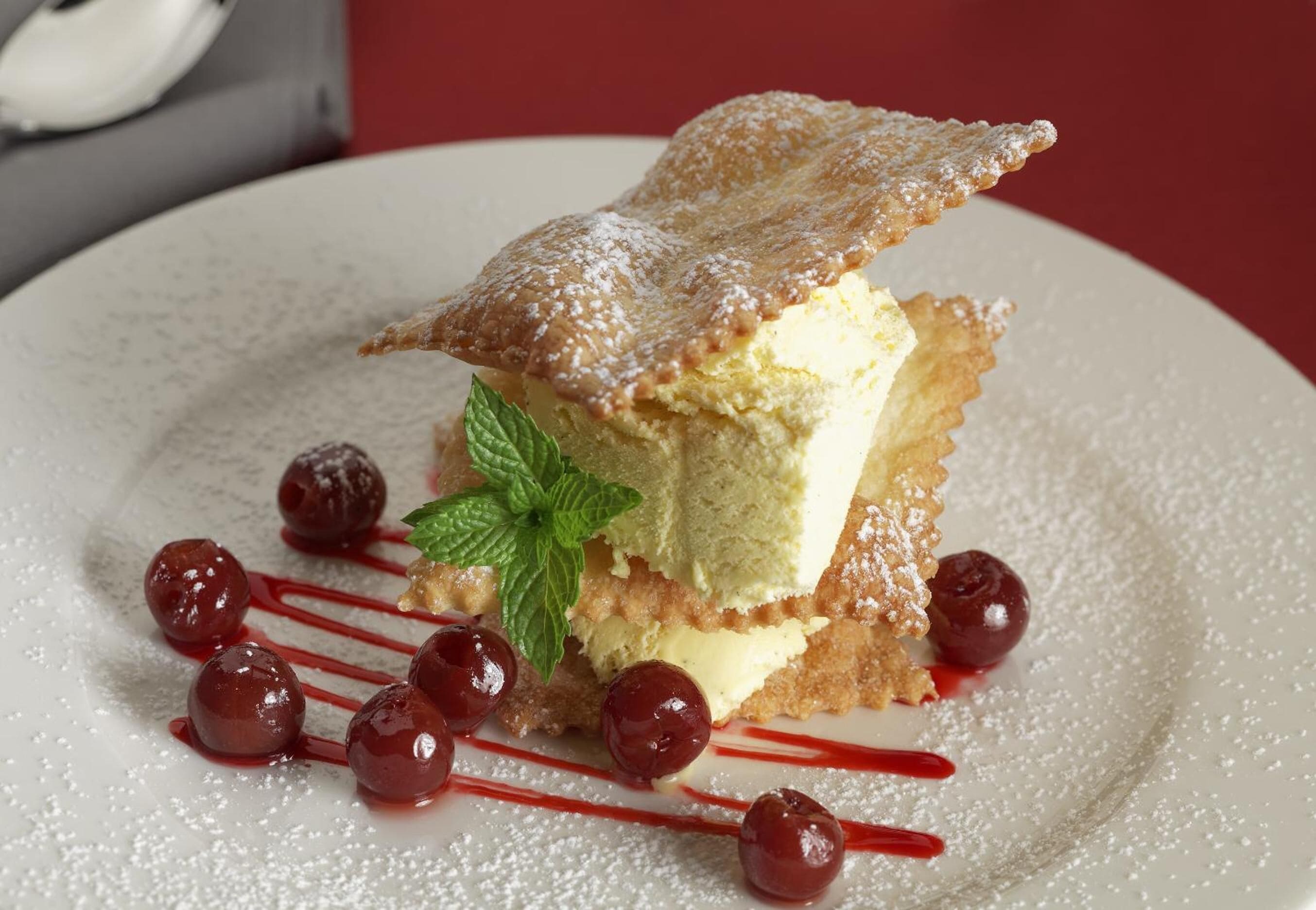 Un postre con un poco de azúcar glas, cerezas y una hoja de menta sobre un plato.