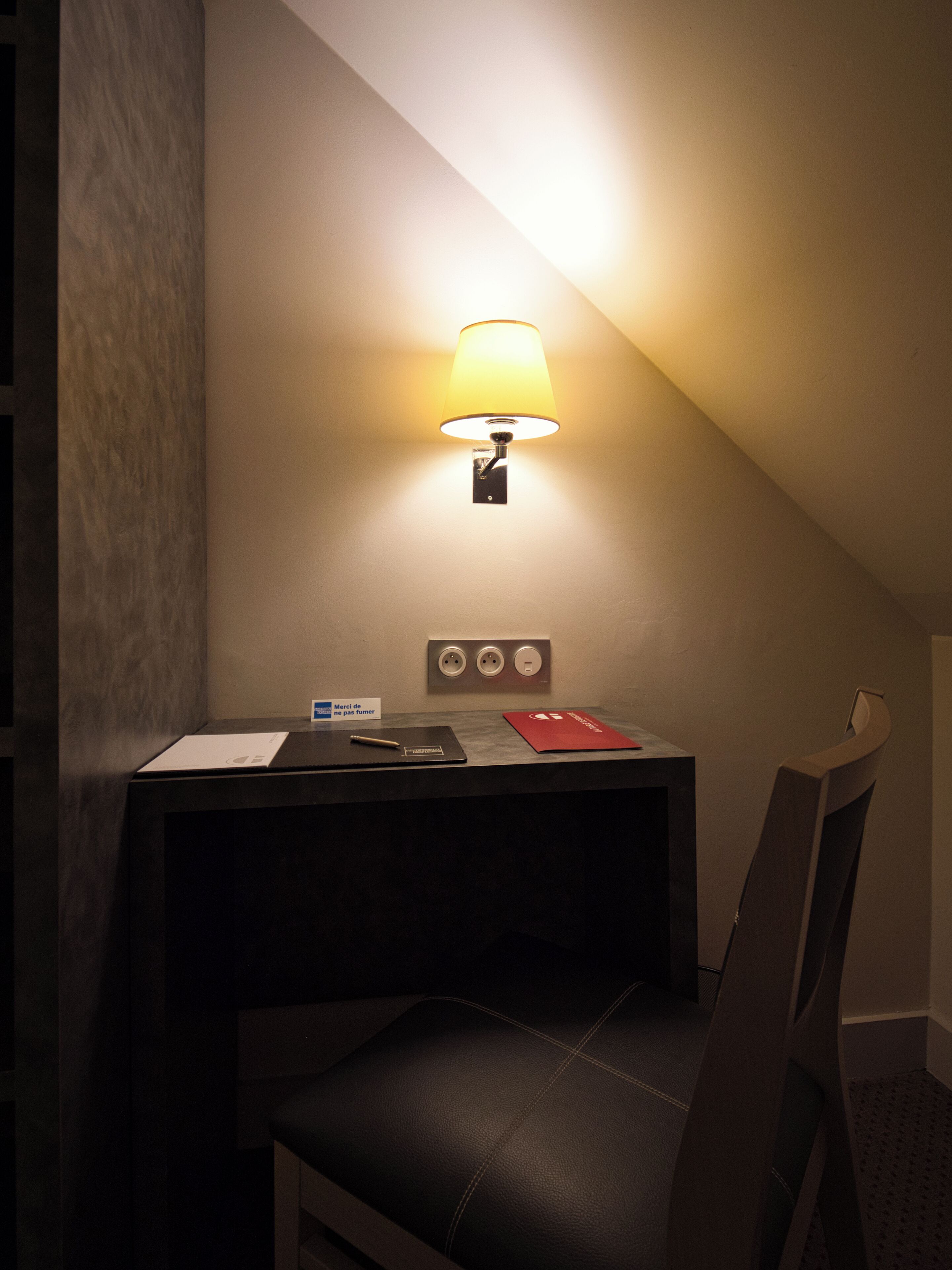 A hotel room with a desk, chair, and a wall-mounted lamp.