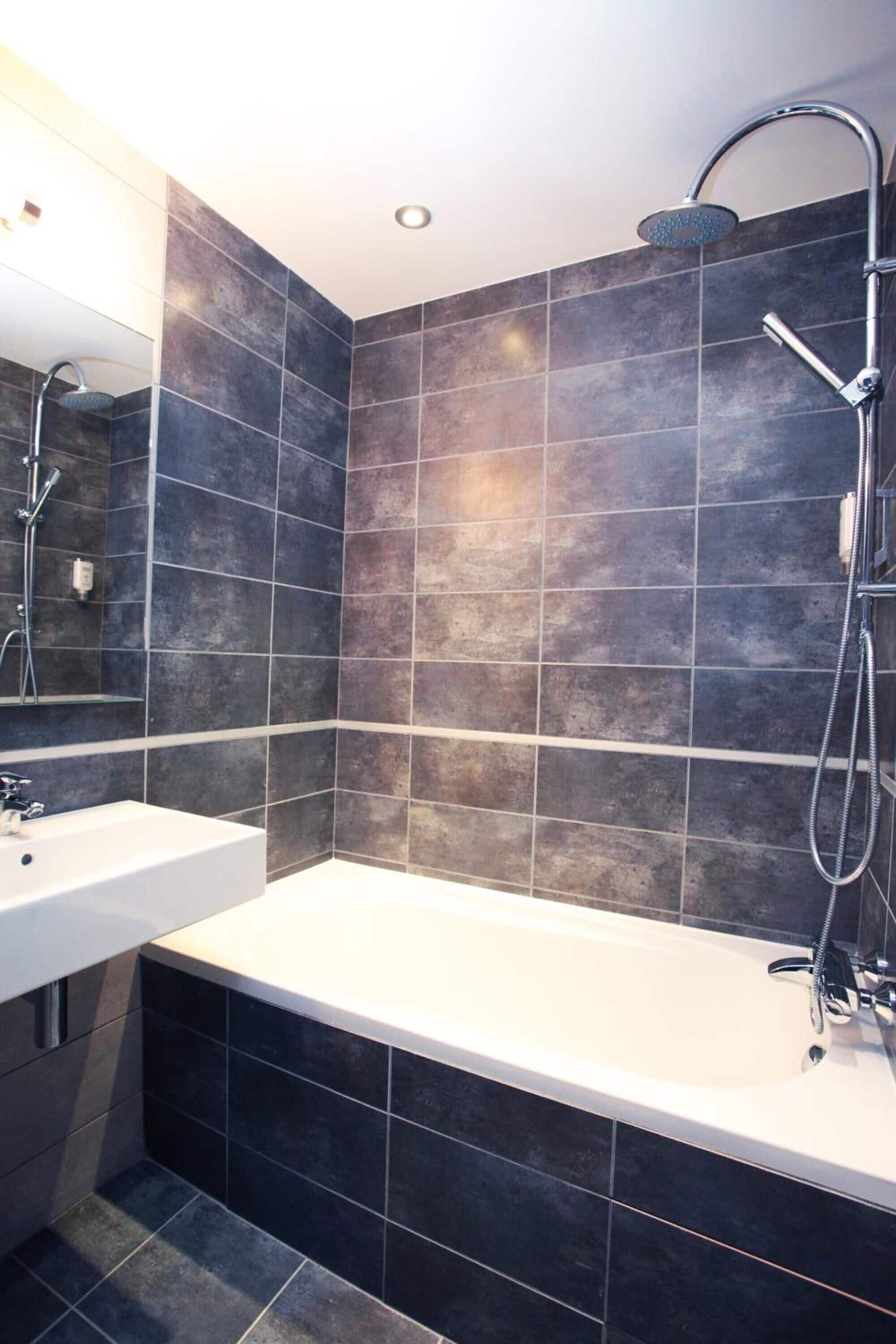 A modern bathroom with dark tiles, a white bathtub, and a handheld showerhead.
