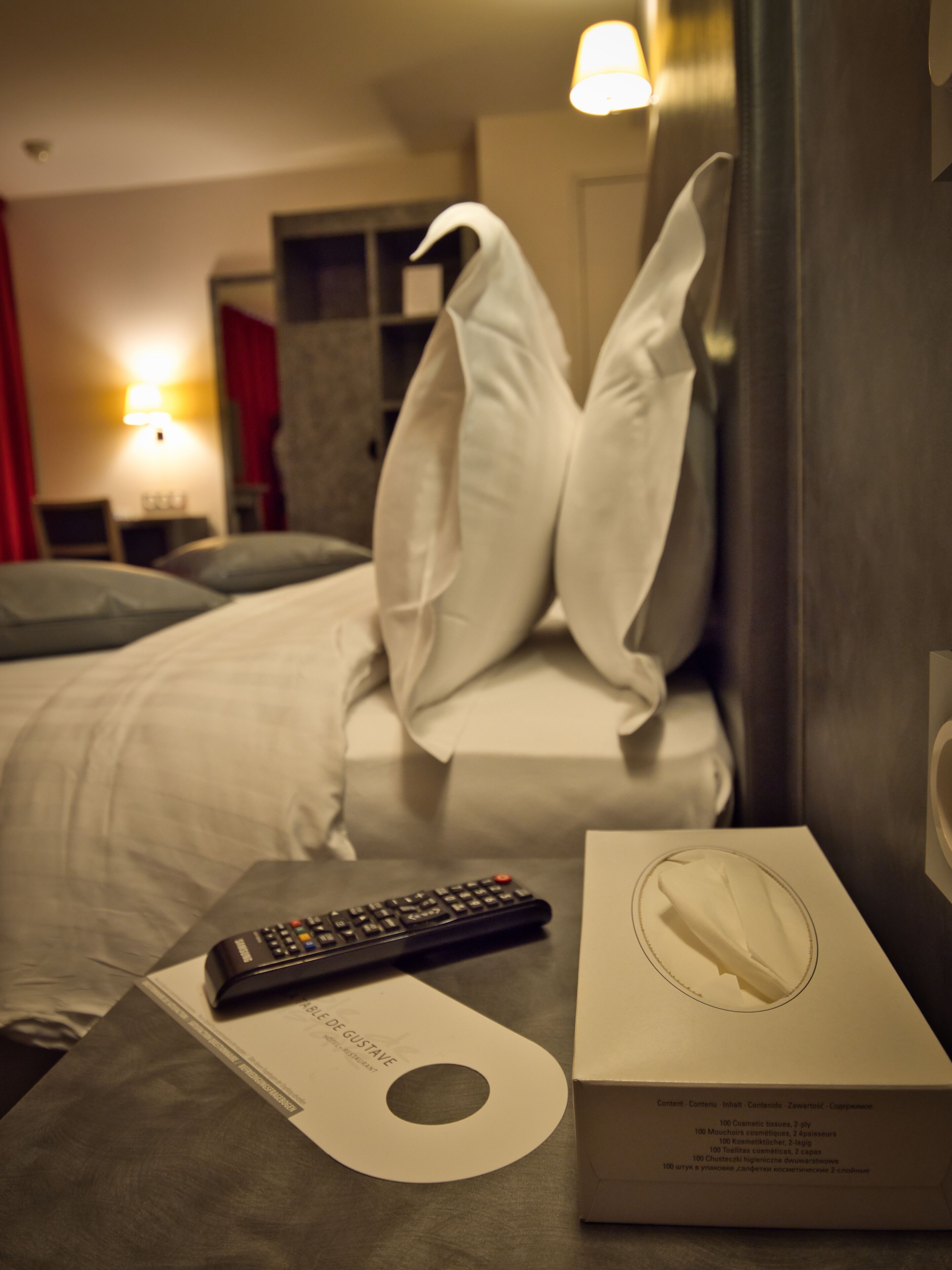 Una habitación de hotel con una cama, almohadas, un mando a distancia y una caja de pañuelos sobre una mesa.