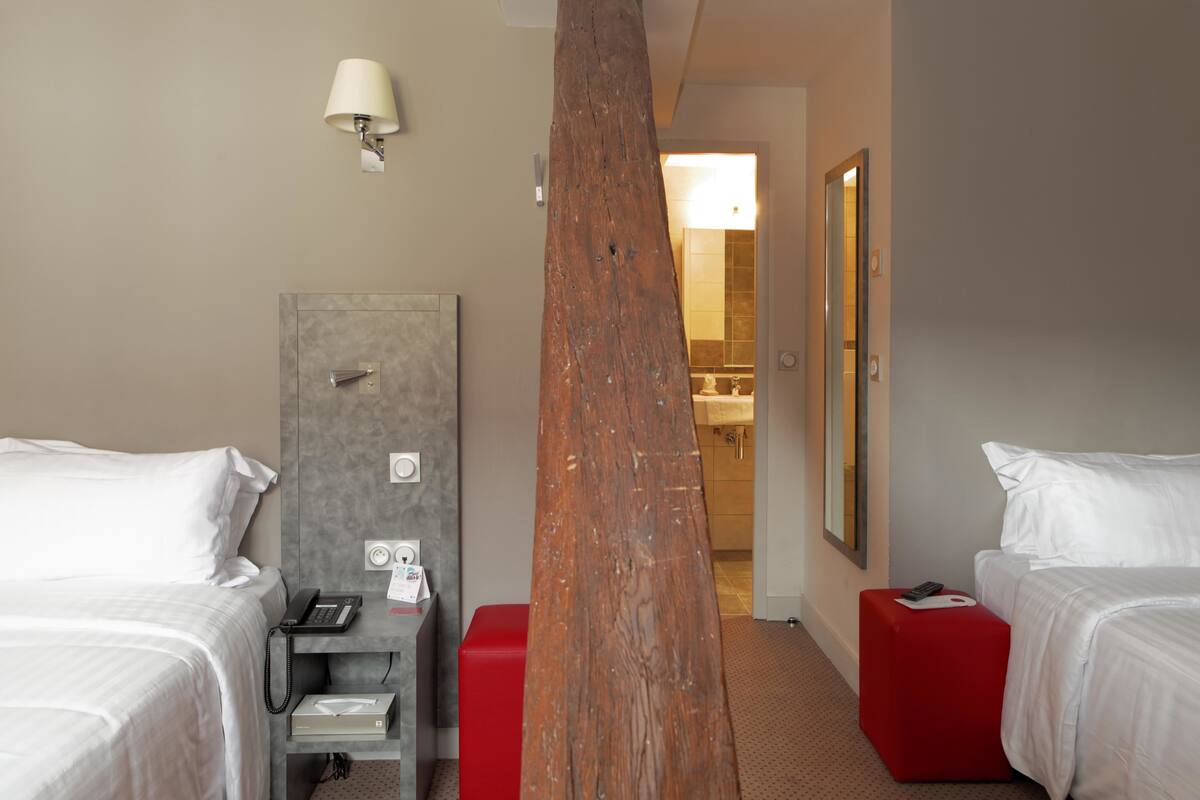 Habitación de hotel con dos camas, un reposapiés rojo, un pilar de madera, un escritorio con teléfono y un espejo en la pared.