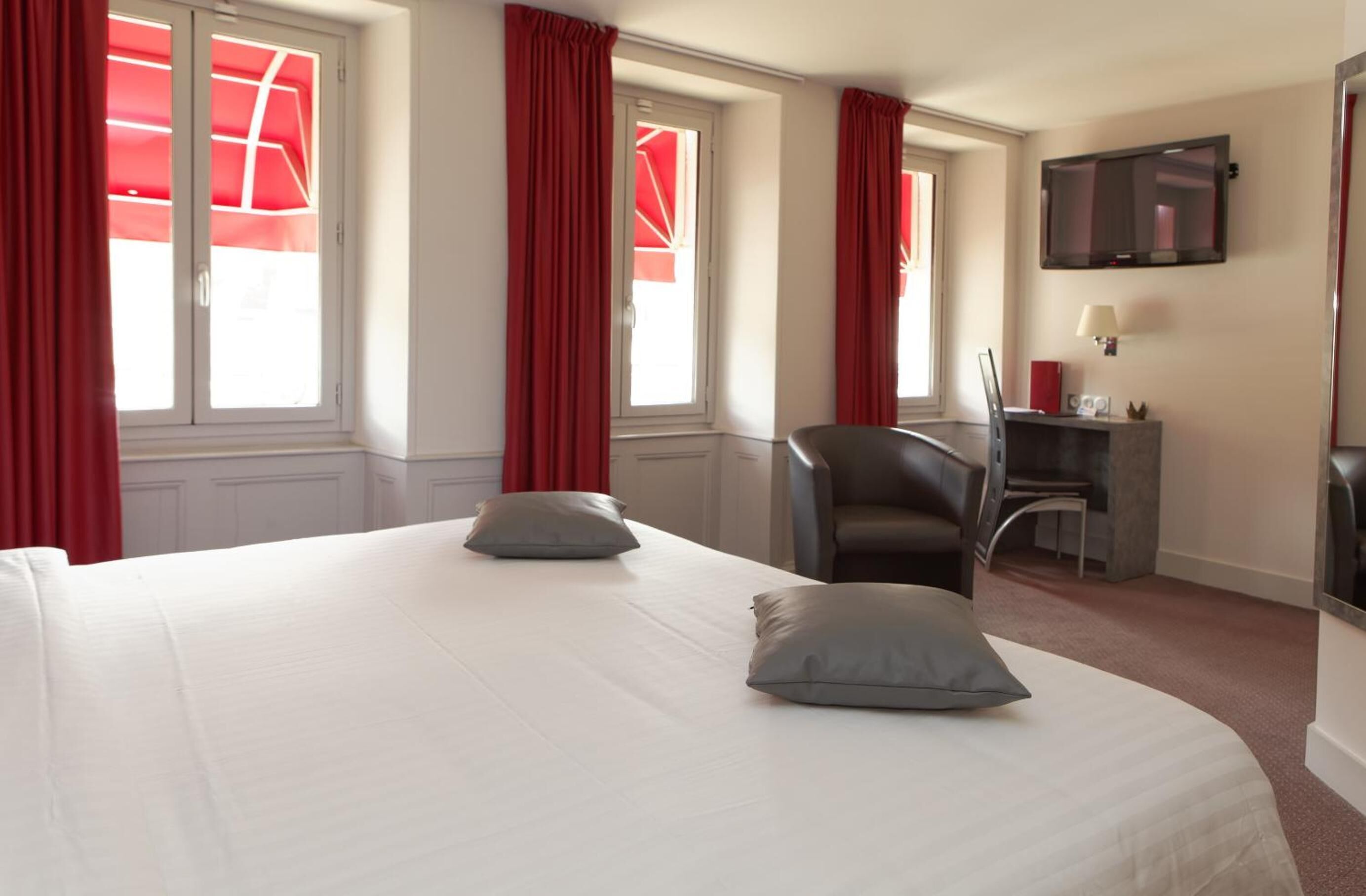 A hotel room with a large bed, two pillows, a desk with a chair, a TV mounted on the wall, and windows with red curtains.