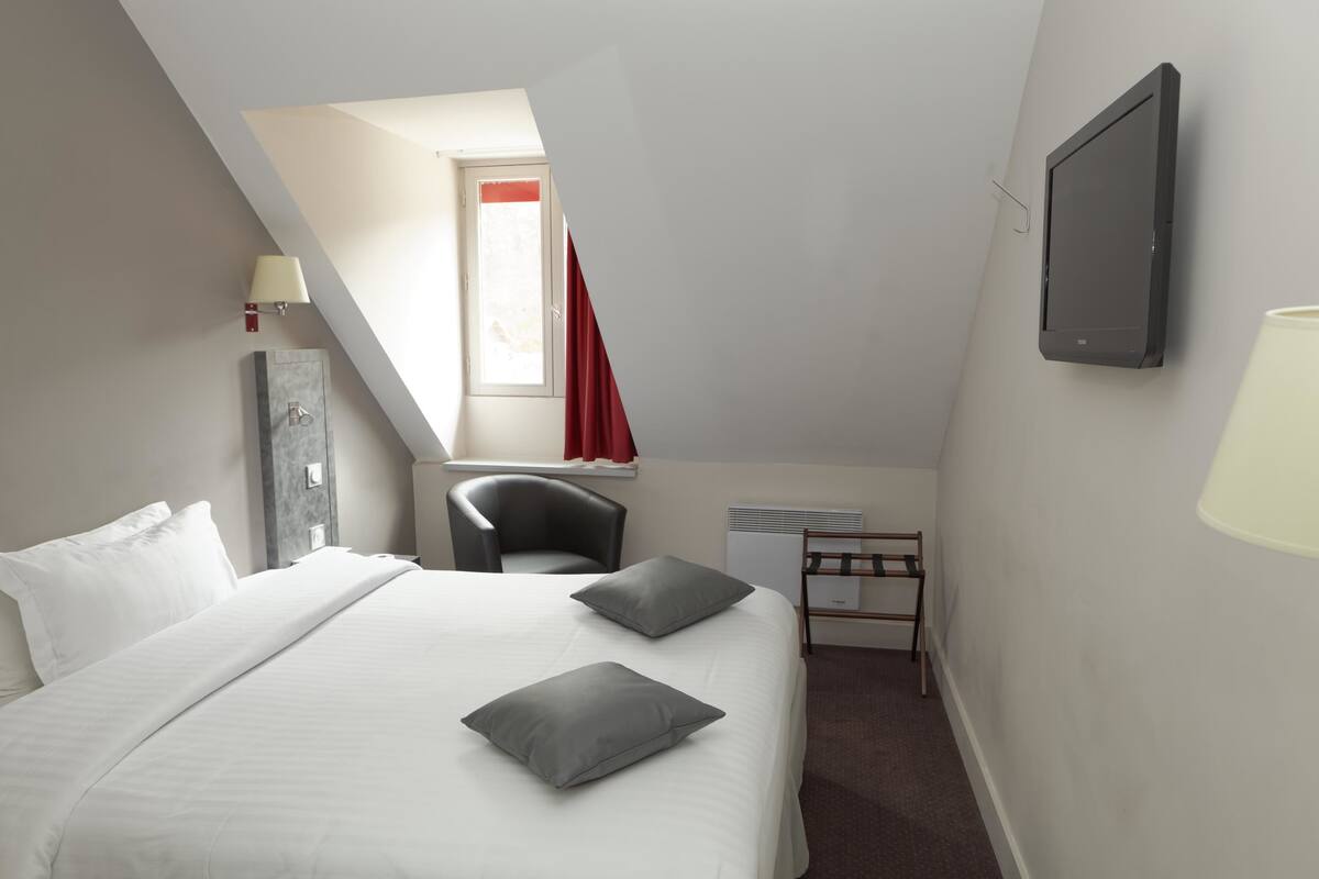 Una habitación de hotel con cama, escritorio, silla, televisor y ventana con cortinas rojas.