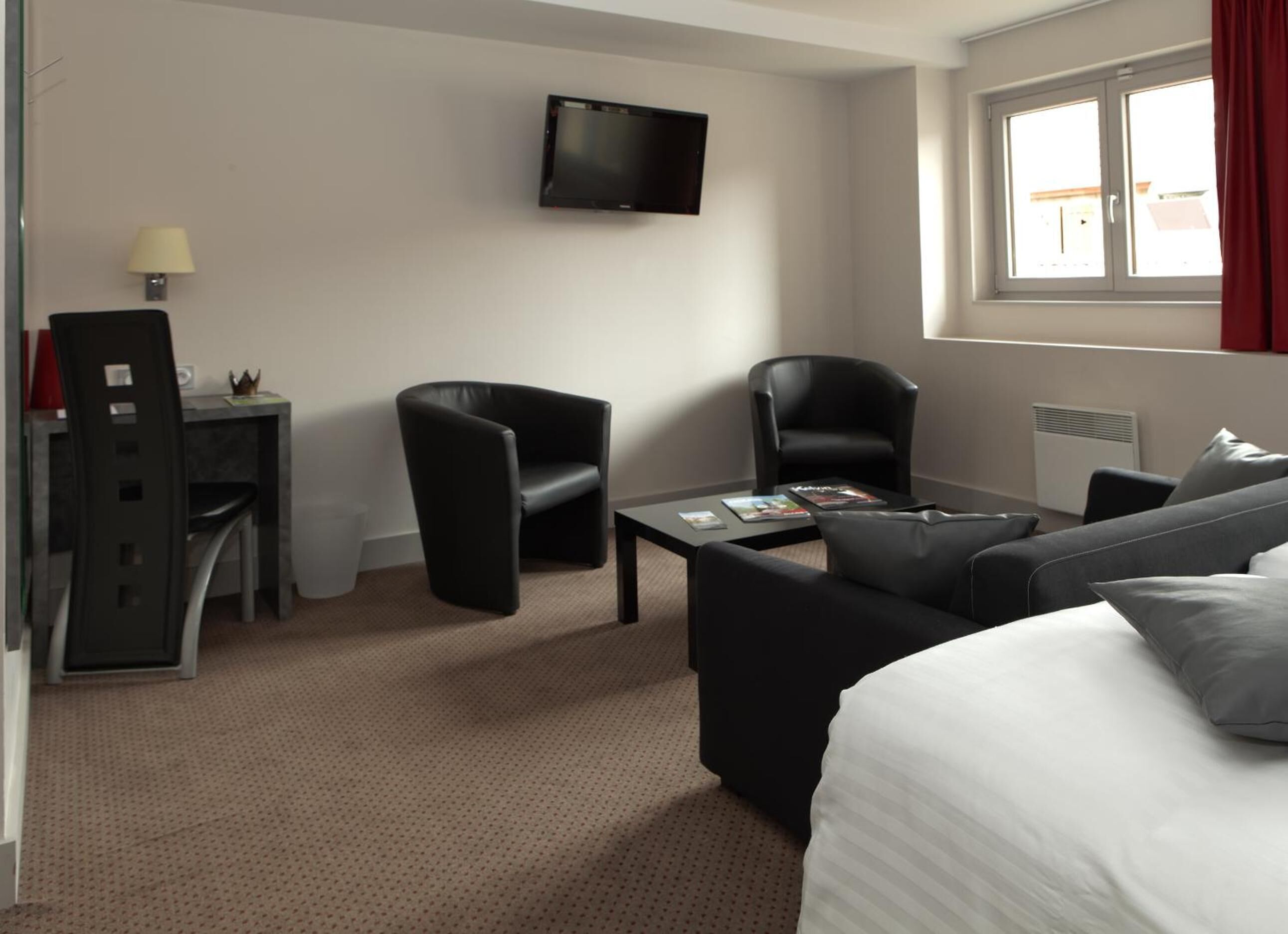A hotel room with a bed, two black armchairs, a small table with magazines, a TV mounted on the wall, and a window with curtains.