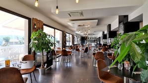 A modern restaurant with high ceilings, pendant lighting, and large windows. Tables are arranged for dining, and there are potted plants adding greenery. - Hotel Perla Riviera (Cagnes-sur-Mer)