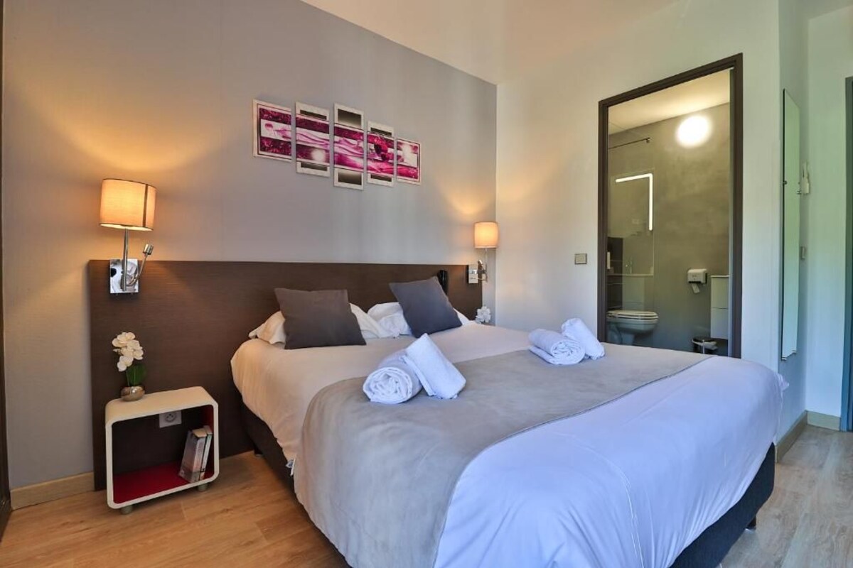 Una habitación de hotel con una cama grande, lámparas de noche, una mesita de noche con un florero y una puerta que da a un baño.