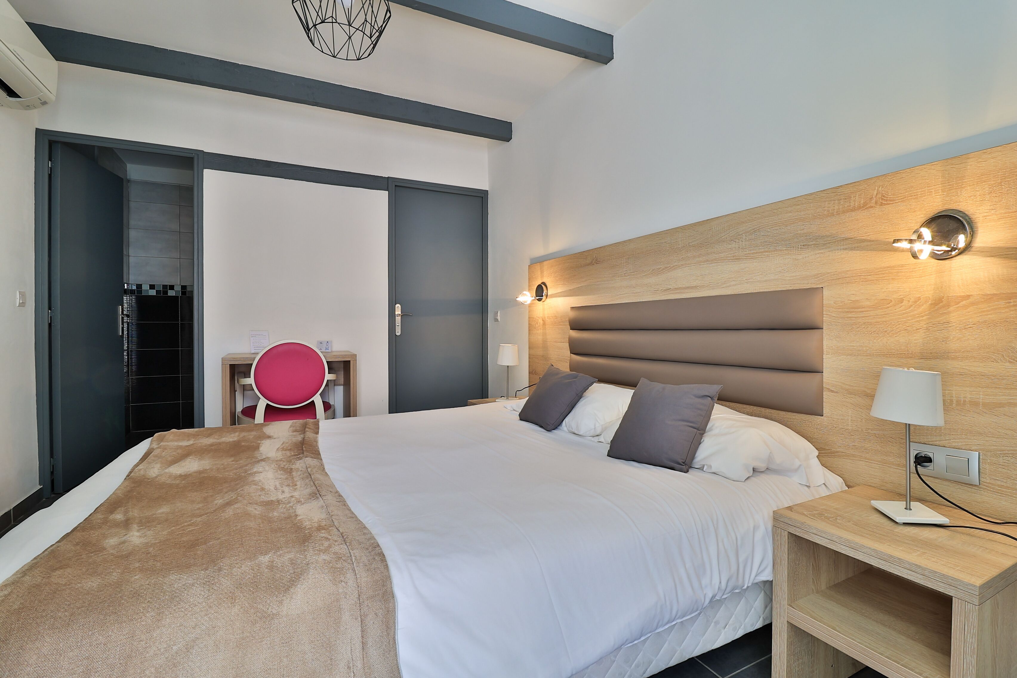 Una habitación de hotel moderna con una cama grande, un cabecero de madera, una mesita de noche con lámpara y un escritorio con silla.
