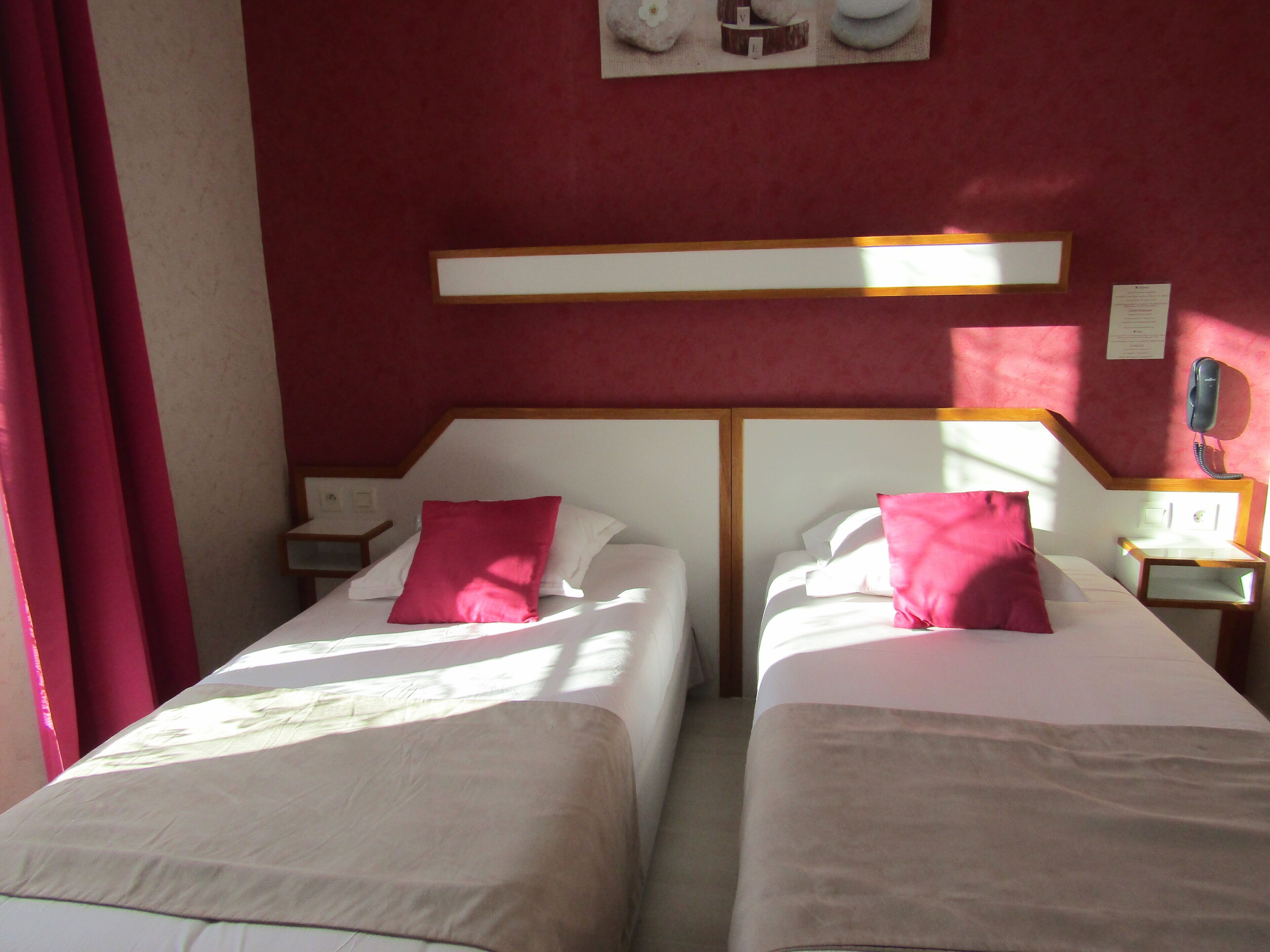 A hotel room with two beds, each with a white headboard and a pink pillow. There is a wooden nightstand with a telephone on each side of the beds.