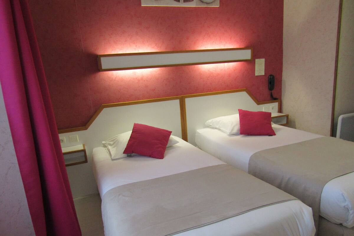 Two beds with white linens and red pillows, a nightstand with a phone, and a wall-mounted light fixture.
