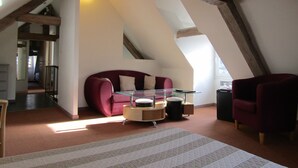 A loft-style room with a red sofa, a glass coffee table, and a red armchair. There is a wooden ceiling with exposed beams and a skylight. - Hôtel Fifi Moulin (Serres)