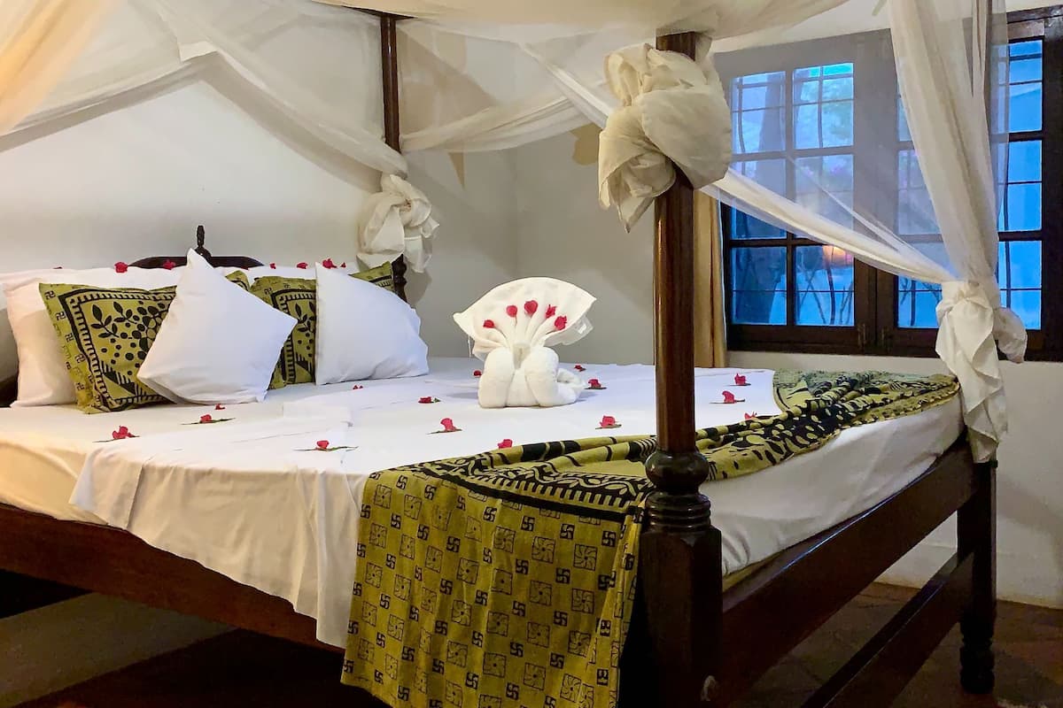 Una cama con dosel, ropa de cama blanca, almohadas estampadas y un arreglo floral decorativo.