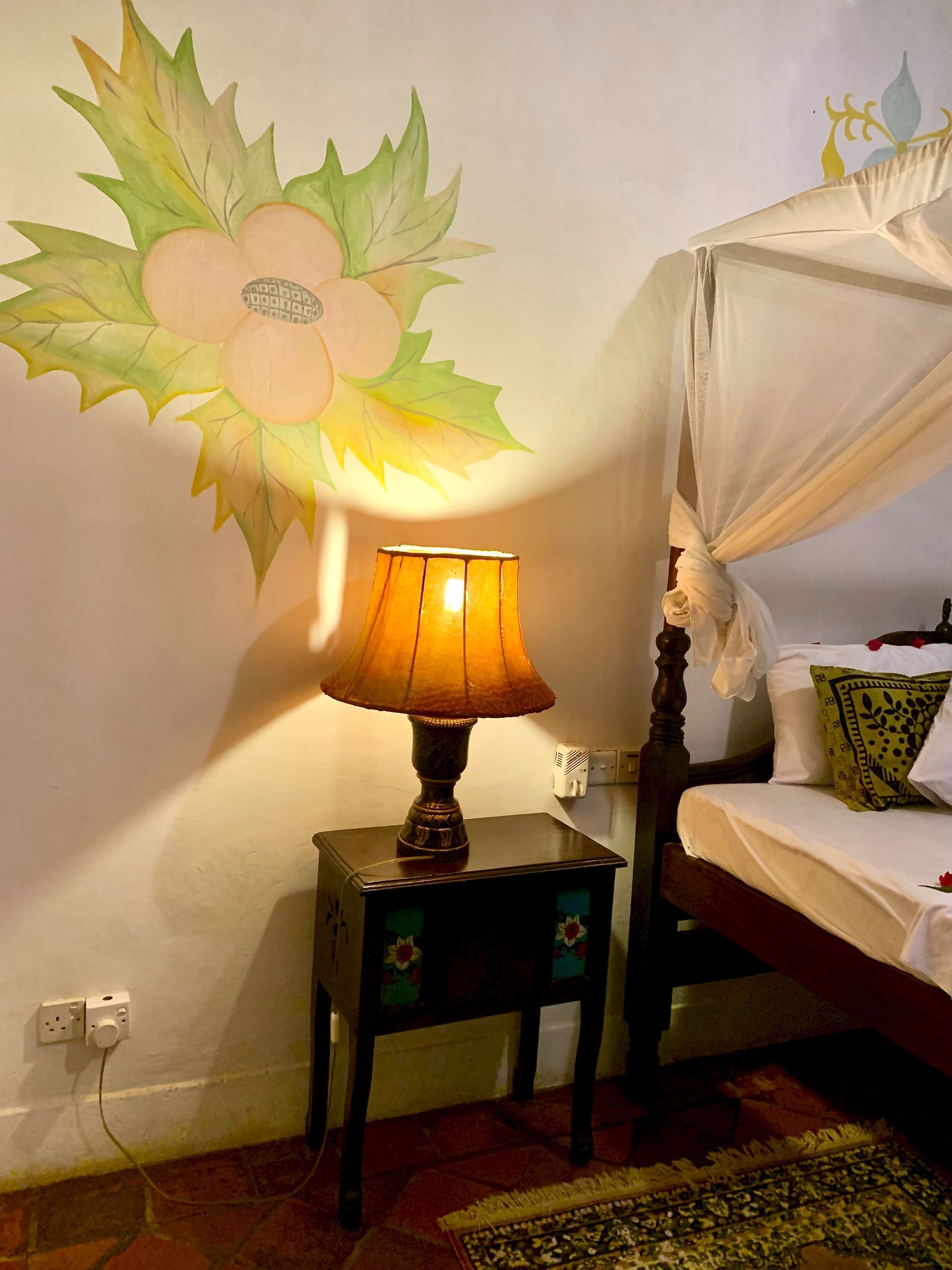 Una cama con dosel, una mesita de noche con lámpara y una pared con un mural floral.