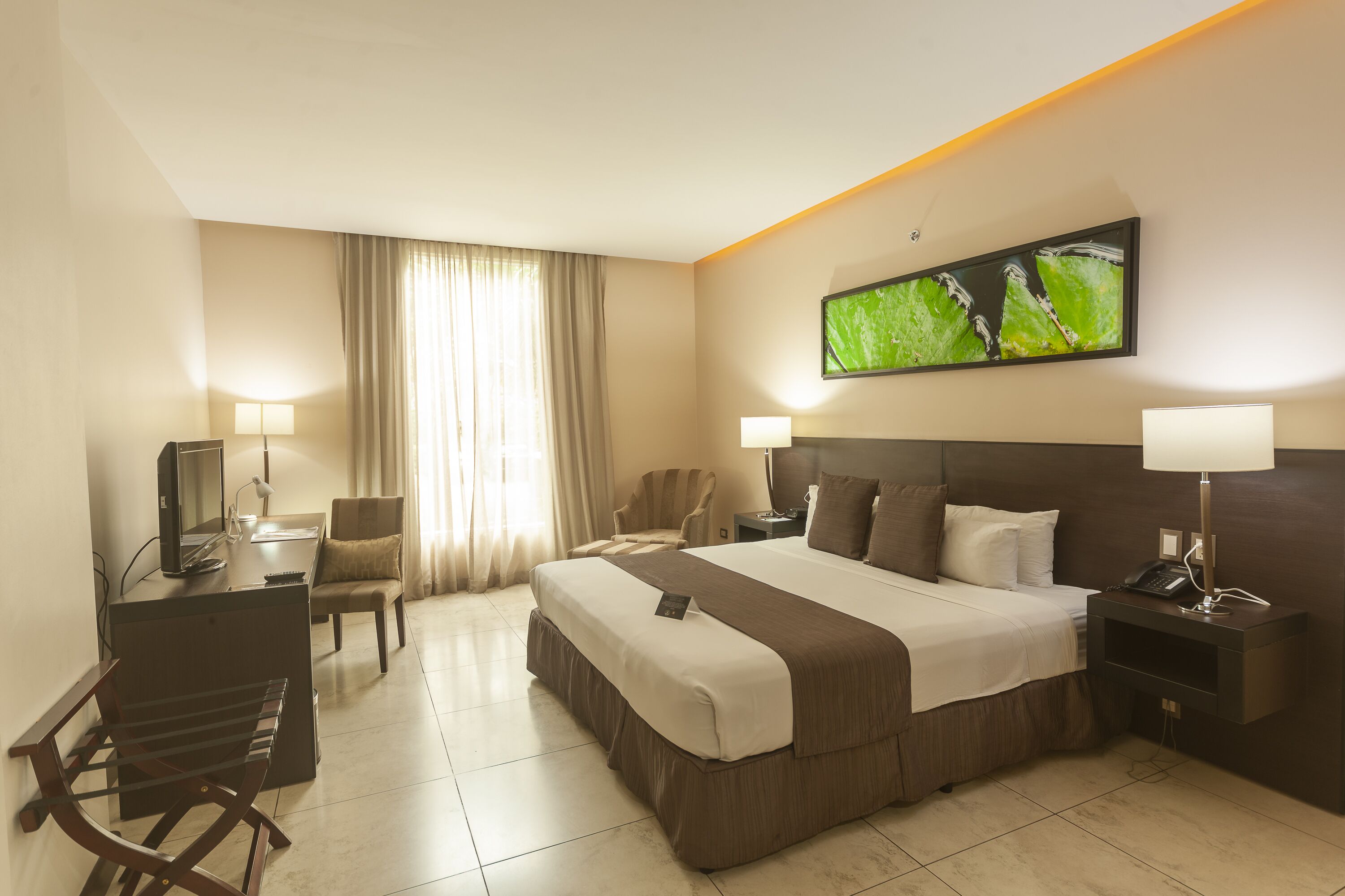 A hotel room with a large bed, two bedside tables, a desk with a chair, a TV, and a window with curtains.